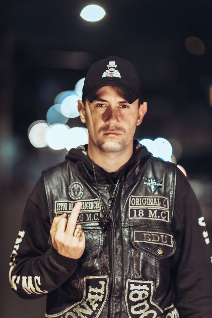ours-journey A man at night wearing a leather jacket and hat, making a defiant hand gesture.