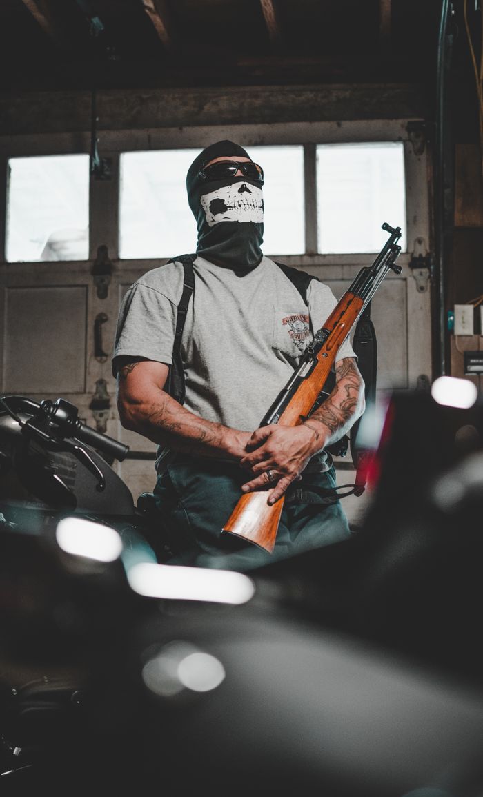 our-experience A masked person stands in a garage holding a rifle, surrounded by motorcycles.